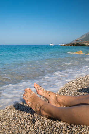 Beautiful female legs on a pebble beach relaxing in blue sea wavesの写真素材