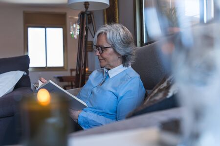 Side photo of an older woman with glasses reading a book sitting on a grey sofa inside a house