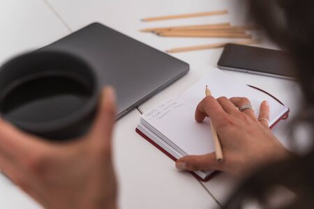 Selective focus on a girl's hand with a pencil in front of a notebook while she holds a coffee sitting in a table with a laptop and a mobile. Teleworking conceptの写真素材