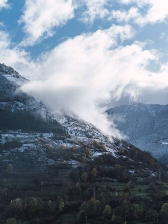 Vertical photo of a snowy mountain hill with trees and the clouds coming off the mountain peak on a sunny dayの写真素材