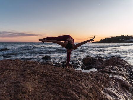 Young woman in sportswear practicing the yoga pose Fire hydrant on some rocks in front of the sea...