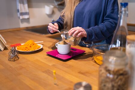 Woman weighing brown sugar on a pink precision scale in the kitchen with other ingredients and...