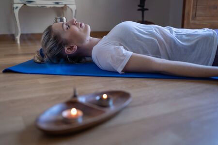 Young woman relaxing on the floor on a blue mat with incense and candles out of focus in the foregroundの写真素材