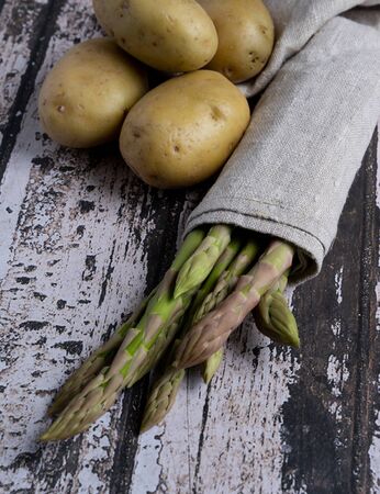 Set of asparagus wrapped in a grey napkin with several potatoes on a worn wooden baseの写真素材