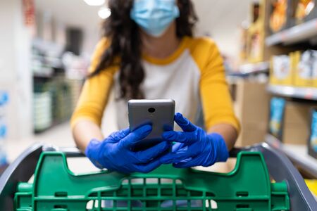 Close-up of a woman leaning on a shopping cart with gloves and a mask consulting her mobile phone in a supermarket aisleの写真素材