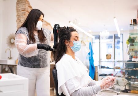 Protected young woman with gloves and mask at the hairdresser consulting current news on her mobile phone while her hair is fixedの写真素材