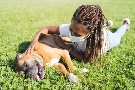 African woman with braids and protective mask stretched out on the grass and stroking a boxer dog in...
