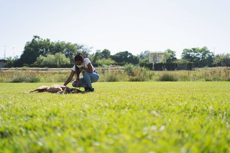 African woman with a mask on her face and braids kneeling on the grass caressing a boxer dog that...