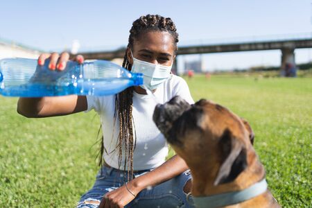 African woman with a mask on her face giving a plastic bottle to a boxer dog on the grassの写真素材