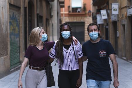 Friends together laughing as they walk down the street wearing a medical mask to prevent the spread of the Covid-19 infection. They are happy for their reunion. Friends with a mask during the new normal.の写真素材