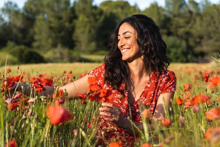 Young brunette woman in a red dress with prints crouched in the middle of a field of poppies touching the flowers while smilingの写真素材