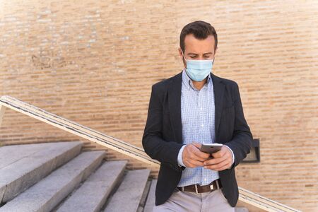 Businessman with a blue jacket and a mask on his face leaning against a railing while using his mobileの写真素材