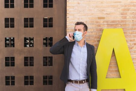 Young businessman formally dressed and with a mask on his face talking to his mobile phone in front of a metal door with a letter next to itの写真素材