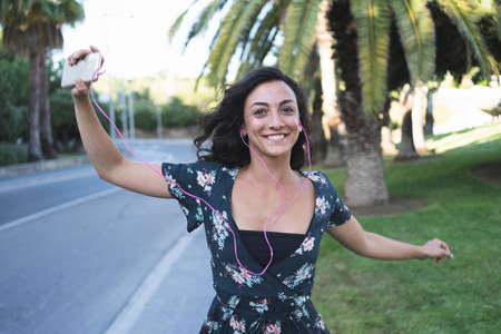 Woman in blue dress dancing by the side of a road while listening to music on her mobile phone with...