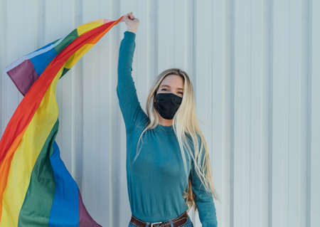 Blonde girl with white background and mask proudly waving the LGBT flag. Concept of pride,...