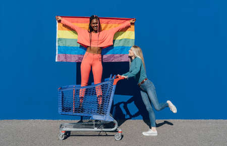 Two proud multiracial friends displaying and claiming LGBT flag on top of a car. Concept of pride,...