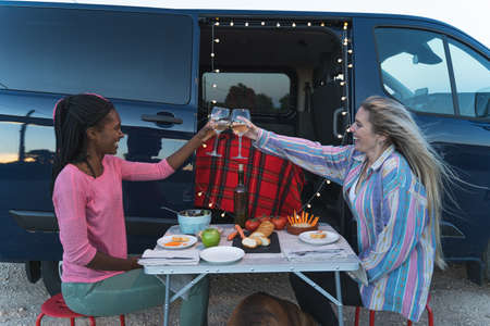 Two woman of different ethnicities toasting with wine at a camping table next to a van parked in the...
