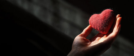 Hand holding a red heart in a beam of light symbolizing love and care, a symbol of affection, kindness, and support on a dark background, a gesture of tendernessの素材