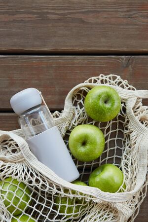 A closeup of an eco bag with green apples and a bottle of water on a wooden surfaceの写真素材