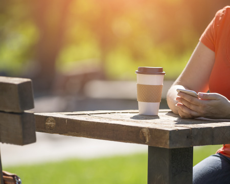 Woman at the park with take away coffee and smart phone in bright sunlightの写真素材