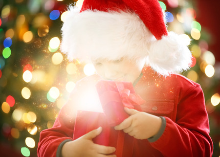Little boy dressed in santa's red hat opening magical gift box in front of decorated Christmas treeの写真素材