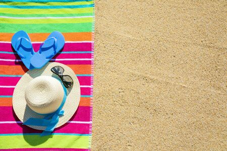 Beach towel with hat, sunglasses and flip flops photographed from above on sandy beach, hot summer day accessories, vacation destination, sunny tropical beach backgroundの写真素材