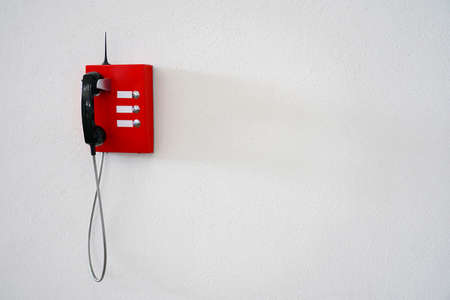 A red phone with a black handset and an antenna hangs on a white wall. Phone number for calling...