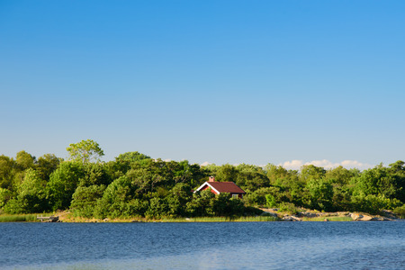 A lonley old red hut on an island in the archipelago. Embedded by green trees. Water in front. Blue sky. Stockholm, Sweden.の写真素材