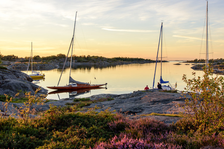 Sunset light over sailboats moored at small rocky island in the outer part of the archipelago. Old wooden sailboat in focus. Stockholm, Sweden.の写真素材