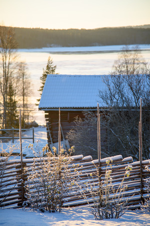 Rural winter scene. Old fence in front. Wodden brarn, a few trees with a frozen lake behind. Forrest as bakground Low sunlight. Dalarna, Sweden.の写真素材