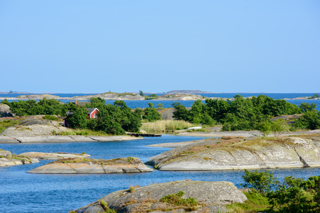 A lonley old red hut on a skerry in the archipelago. Embedded by green trees. Water and low skerries in front and behind. Blue sky. Stockholm, Sweden.の写真素材