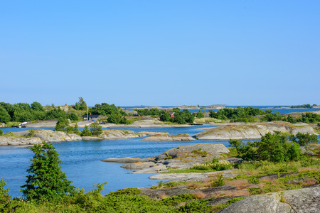 Small huts on a skerry in the archipelago. Embedded by green trees. Water and low skerries in front and behind. Blue sky. Stockholm, Sweden.の写真素材