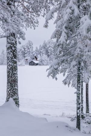 Snowcapped island with cottage and boat house seen from another snowy island. Tree trnuks in front.の写真素材