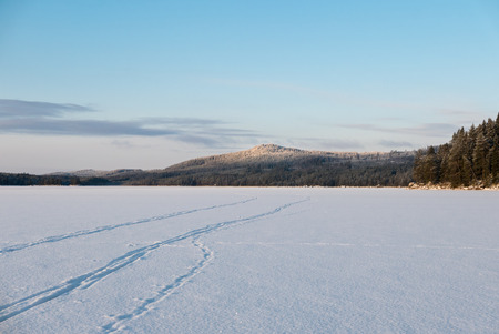 Frosen lake with snow and man made ski tracks. Frosty forest hills at distance. Blue sky.の写真素材
