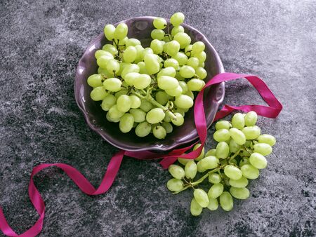 Fresh green grapes on a purple bowl and on a dark grey table with a pink ribbon, top view and selective focusの写真素材