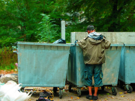 Poor and hungry homeless man in dirty clothes looking for food in the dumpster on the urban street in the city. Social problems of modern society. Lifestyle of tramp, living in the streets.の写真素材