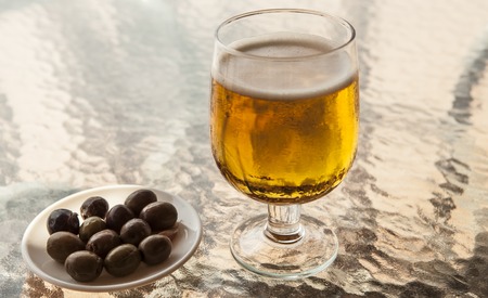 Misted glass of beer with olives on a glass table.の写真素材