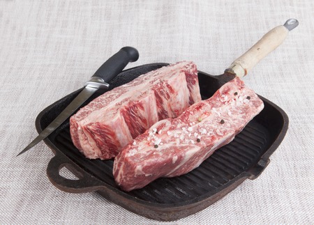 Close-up of two pieces of fresh marbled beef with sea salt and black pepper, knife on a cast-iron grill pan.の写真素材