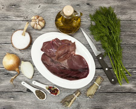 Pieces of fresh raw beef liver, onion, garlic, spices, dill, parsley, salt, knife, olive oil on porcelain plate on a wooden backgroundの写真素材