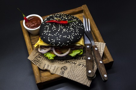 Black burger. A burger with a black roll slices of juicy marble beef, fused cheese, fresh salad and sauce of a barbecue. A burger on the newspaper on a wooden tray with a knife and a fork on a dark backgroundの写真素材