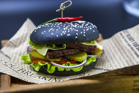 Black burger. A burger with a black roll slices of juicy marble beef, fused cheese, fresh salad and sauce of a barbecue. A burger on the newspaper on a wooden tray  on a dark backgroundの写真素材