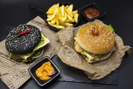 Set of the black burger and classical American burger . Black burger roll slices of juicy marble beef, fused cheese, fresh salad with French fries on a cast-iron griddle, with chili sauce and guakomole, classical juicy burger with chicken and ham on a wooの写真素材