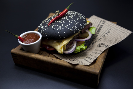 Black burger. A burger with a black roll slices of juicy marble beef, fused cheese, fresh salad and sauce of a barbecue. A burger on the newspaper on a wooden tray with a knife and a fork on a dark backgroundの写真素材