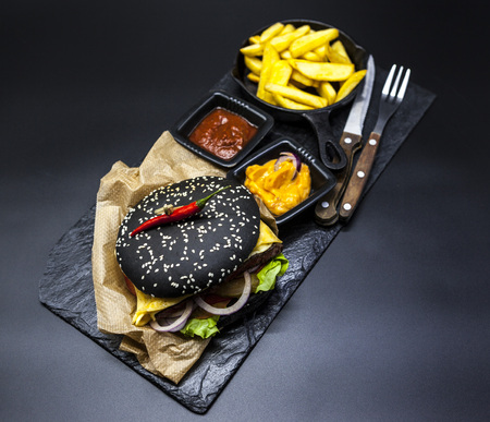 Black burger. Set of the black burger. Stone plate with: black burger roll slices of juicy marble beef, fused cheese, fresh salad with French fries on a cast-iron griddle, with chili sauce and guakomole.の写真素材