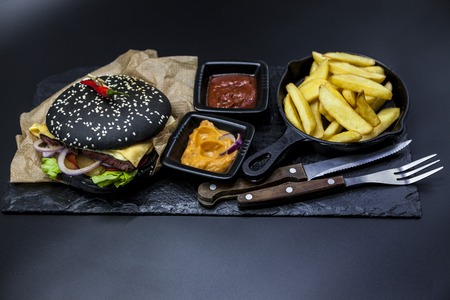 Black burger. Set of the black burger. Stone plate with: black burger roll slices of juicy marble beef, fused cheese, fresh salad with French fries on a cast-iron griddle, with chili sauce and guakomole.の写真素材