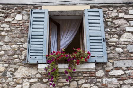 Old window with open shutters with flowers on the window sill on the stone wall. Italian Village.