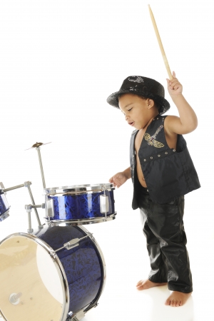 An adorable, barefoot preschooler dressed as a rock star with a drum stick poised high over a drum set, ready to "wham" it.  On a white background.の写真素材