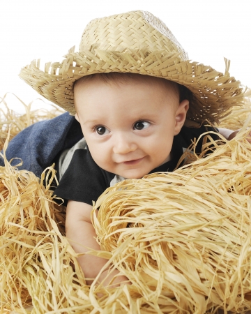 An adorable baby boy wearing a straw hat as he happily lays on a stack of hay  の写真素材