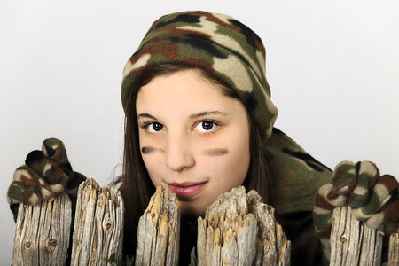 Close-up of an attractive young teen girl in camoflage looking over the top of an old wooden fence. Isolated on gray.  の写真素材