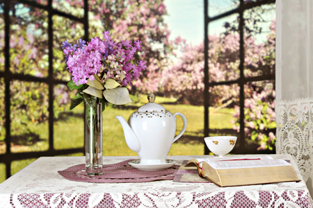 An opened Bible on a table set with a teapot, tea cup and bouquest of violate lilacs, all set before a window opened to a sunny field of lilacs bushes.  Shallow depth of field with focus on the Bible.の写真素材